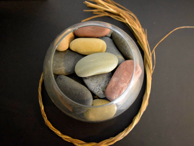 Top view of round glass container filled with stones with a piece of braided sweet grass outside on a black table