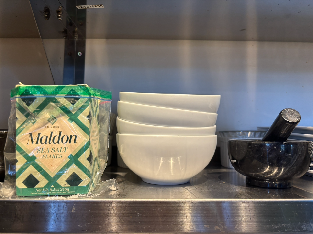 Container of Maldon sea salt sitting on a kitchen shelf next to a stack of bowls